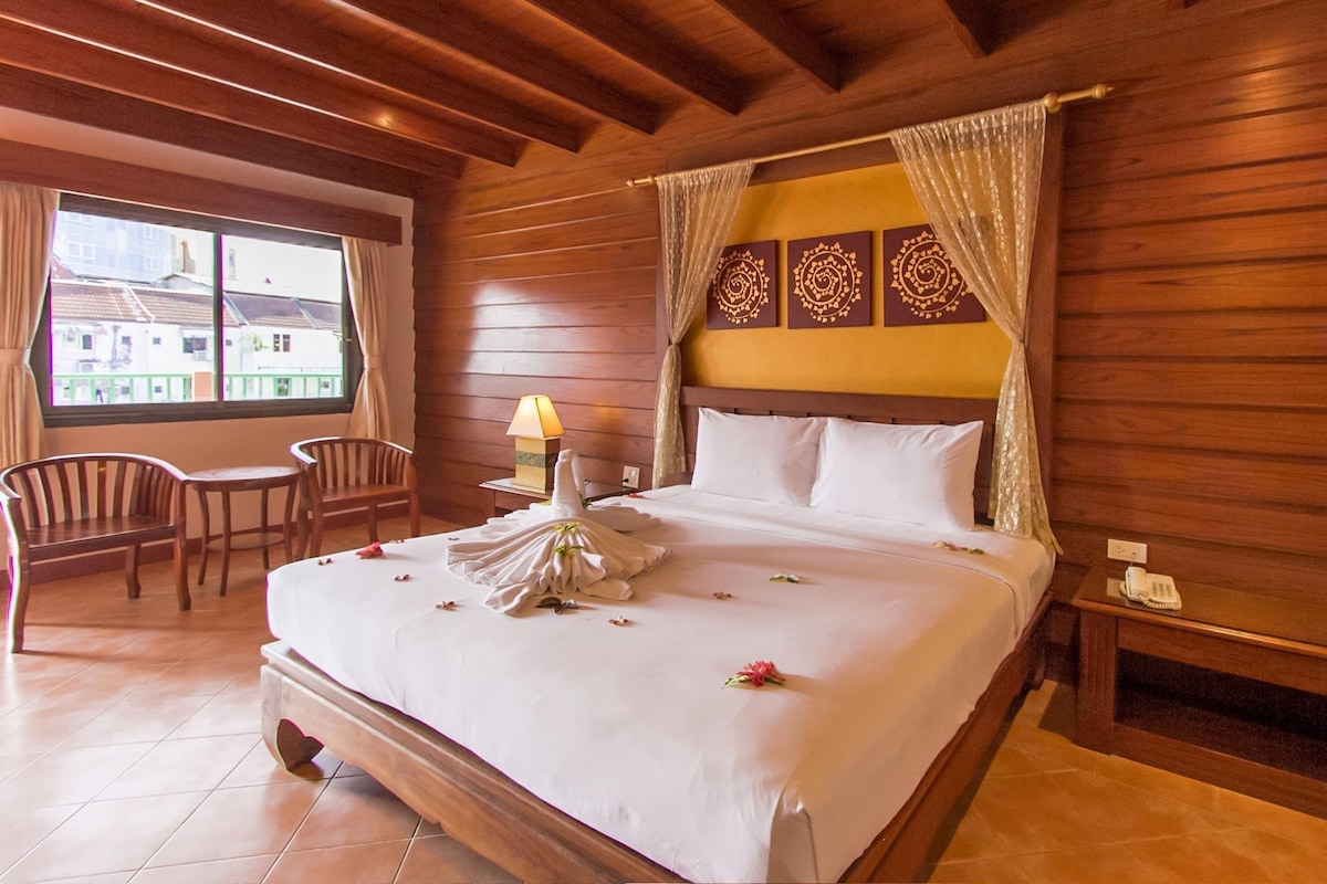 A hotel room with a wooden ceiling, a large bed with white linens, a bedside table with a lamp, a chair, and a window with curtains.