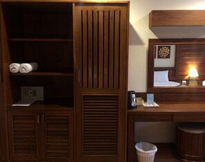 A hotel room with a wooden desk, a bed with a headboard, a nightstand with a lamp, a trash bin, and a wall-mounted heater.