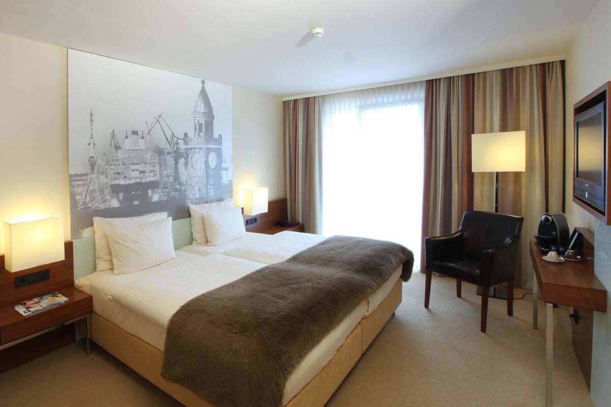 A hotel room with a large bed, a desk with a chair, a TV, and a wall-mounted lamp.