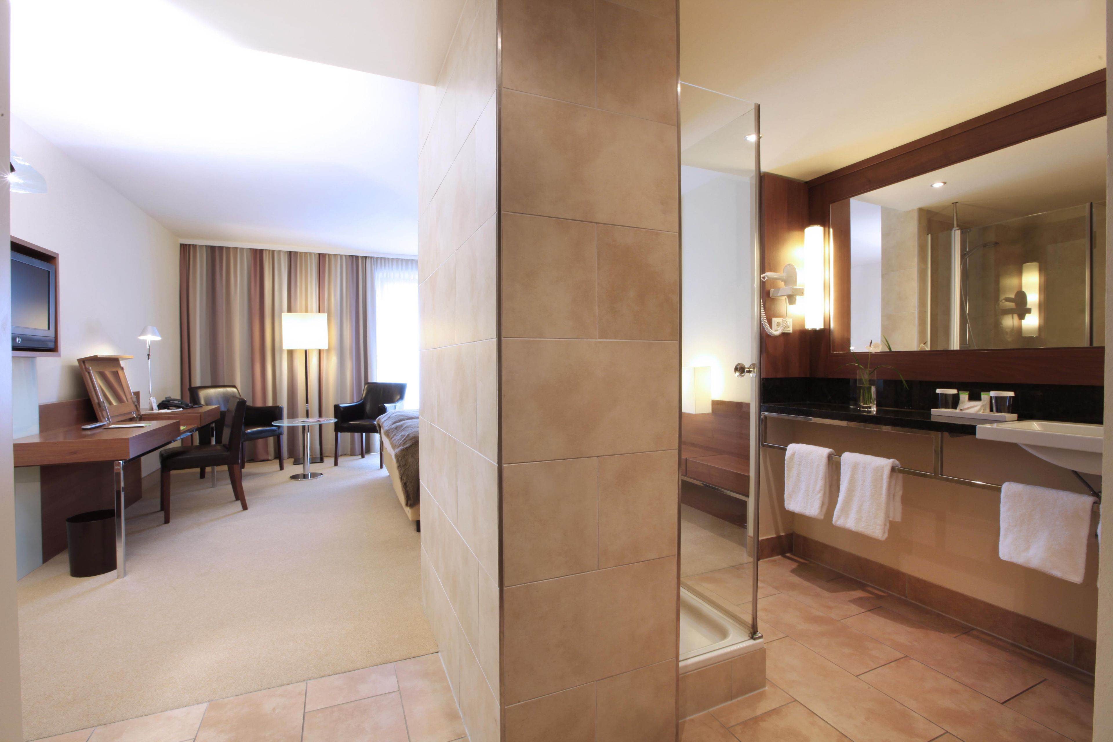 Ein Hotelzimmer mit Schreibtisch, Stühlen, einem Bett, einem Badezimmer mit Dusche und einem großen Spiegel.