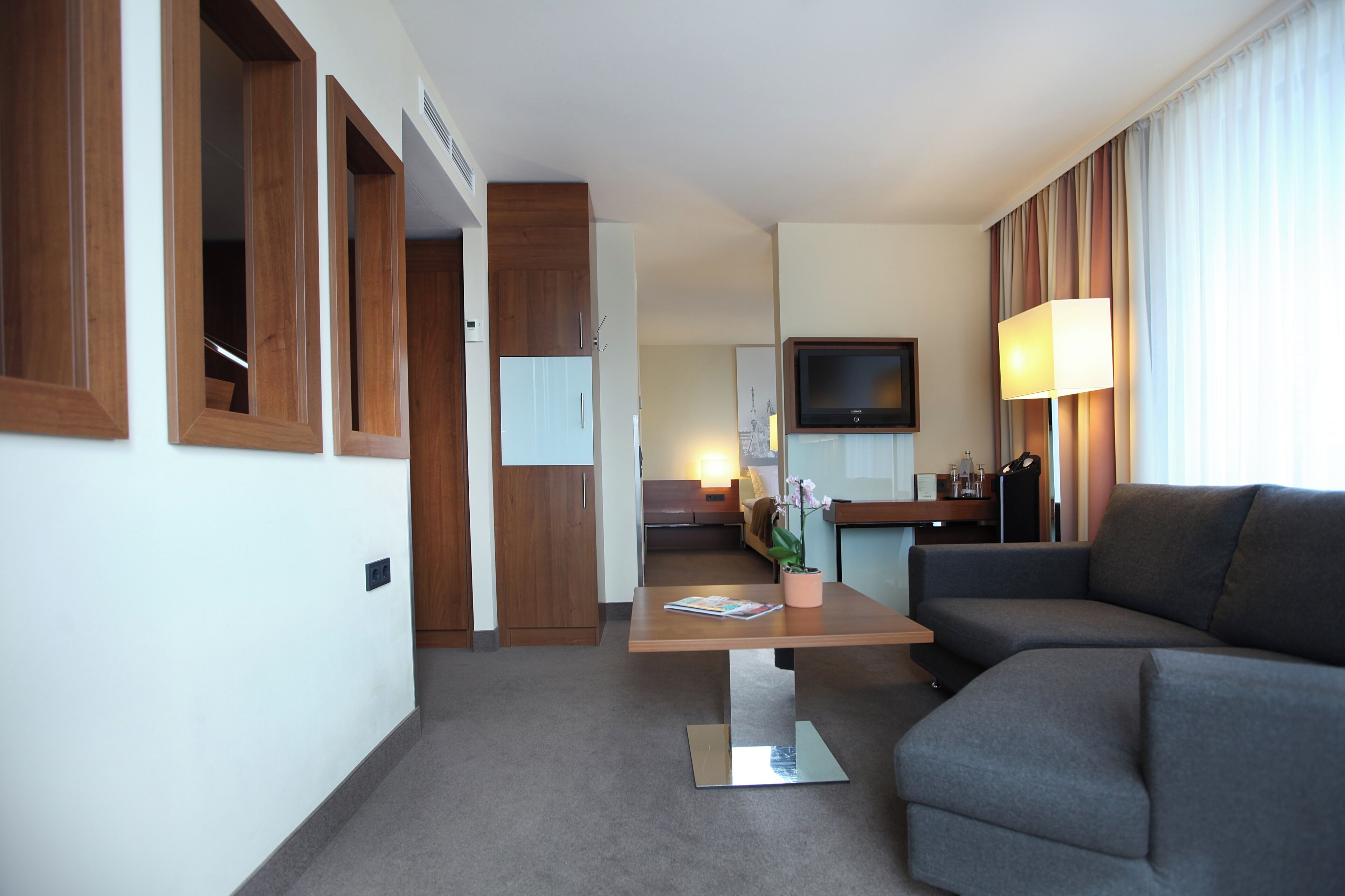 Una habitación de hotel moderna con un sofá gris, una mesa de centro de madera, un televisor de pantalla plana y un amplio ventanal con cortinas.