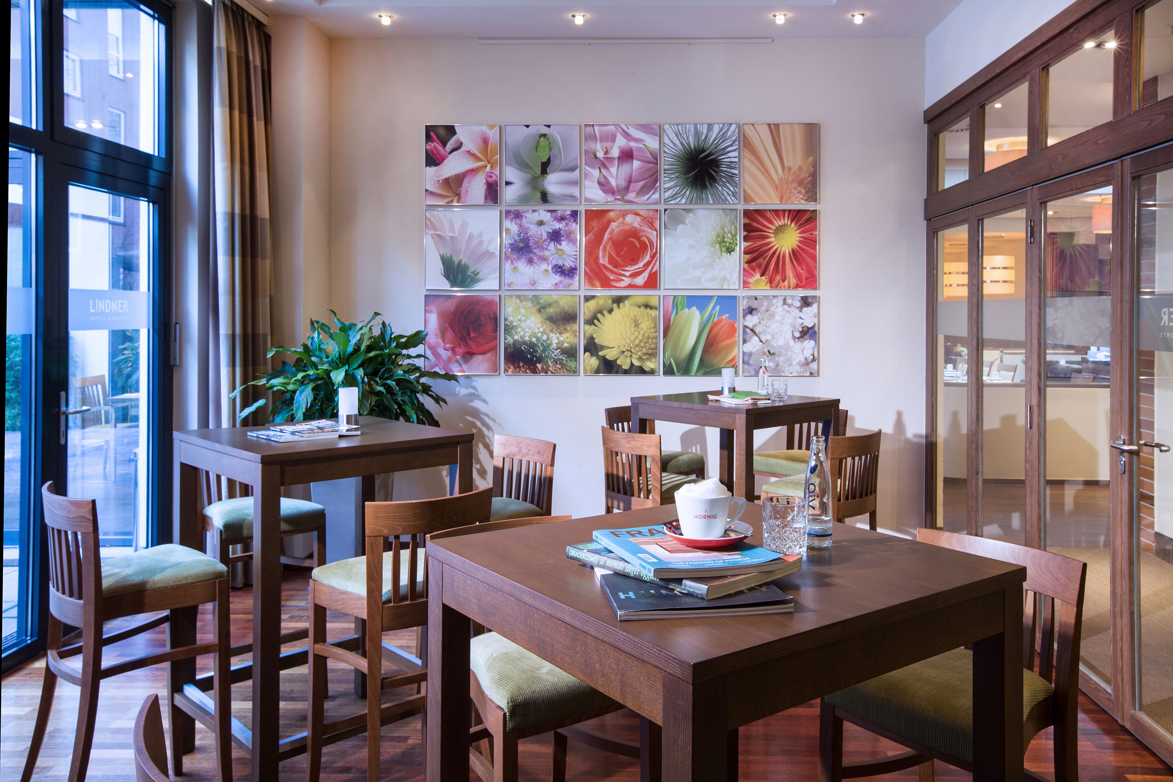 Ein Essbereich mit Holztischen und -stühlen, einem großen Fenster mit Vorhängen und einer Wand, die mit floralen Bildern geschmückt ist.