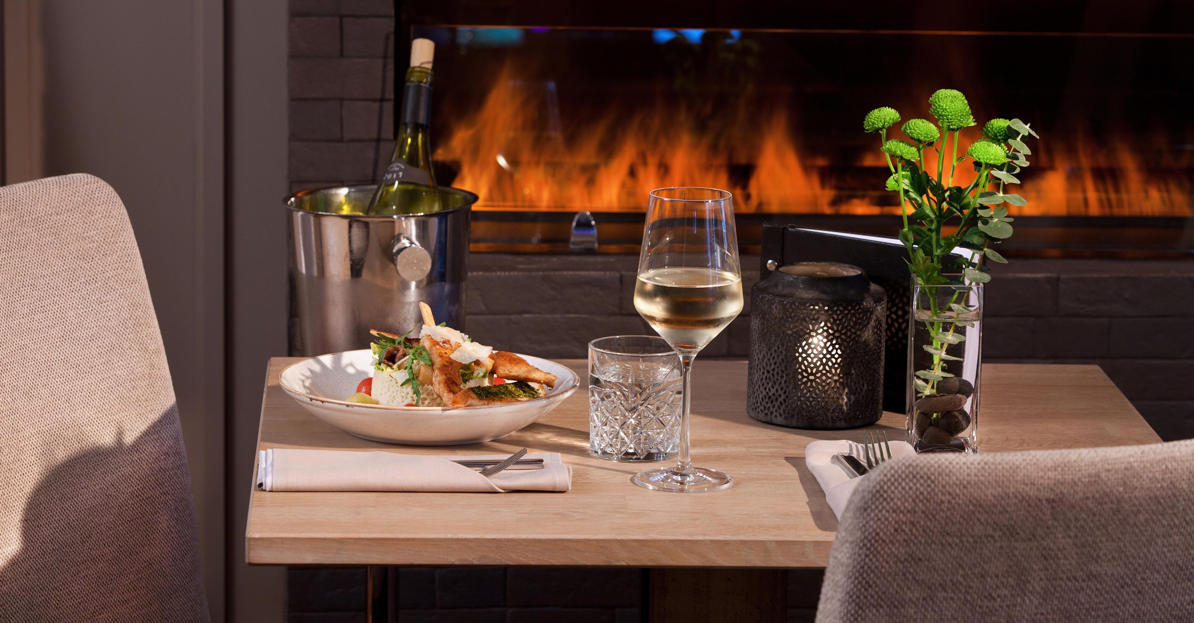 Une table à manger avec une assiette de nourriture, un verre à vin, un verre d’eau et un vase contenant des plantes vertes.