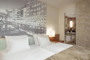 A hotel room with a bed, a nightstand, a lamp, a bathroom with a mirror, and a cityscape mural on the wall.