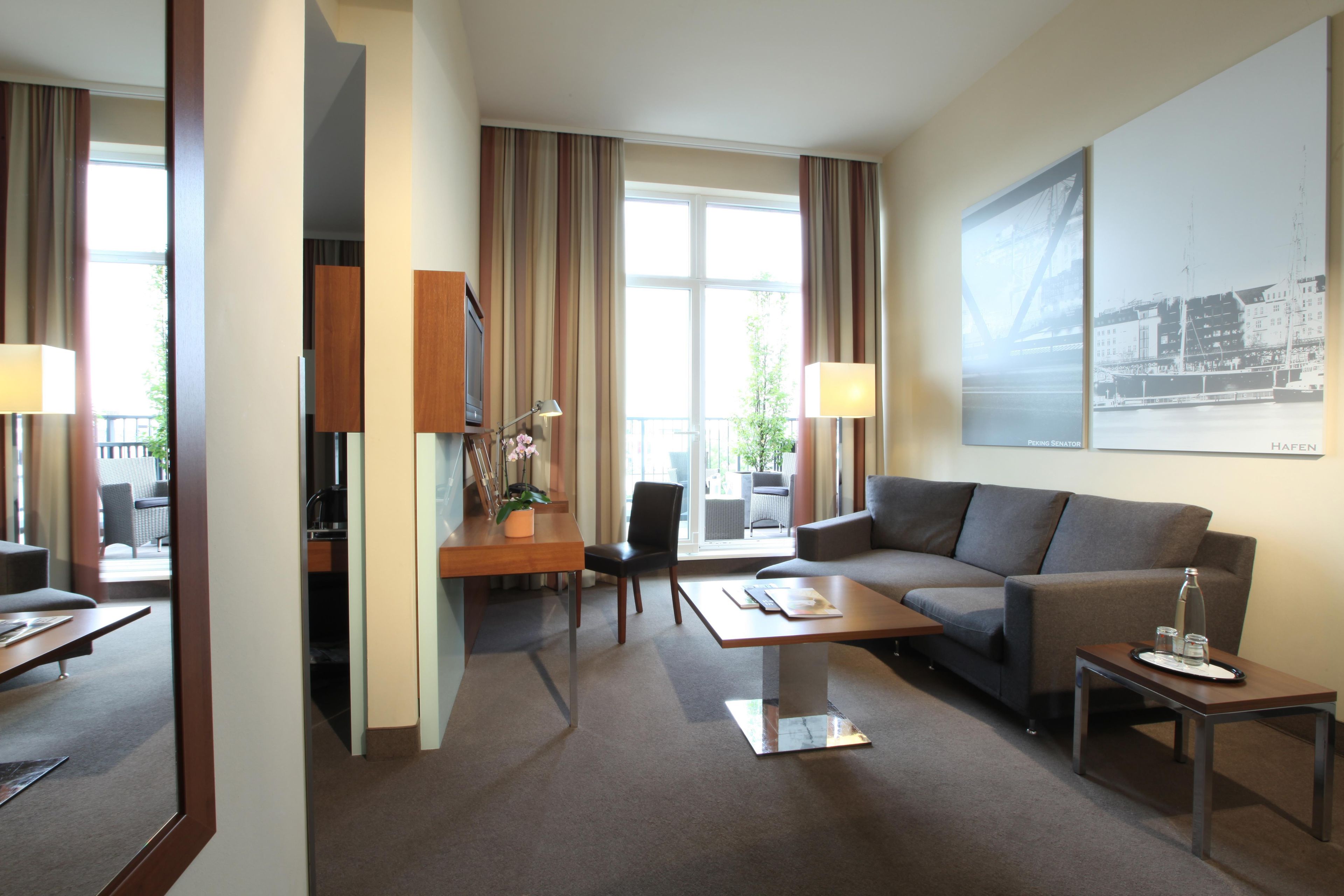 A hotel room with a sofa, a dining table, a chair, a lamp, and a large window with curtains.