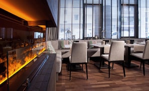 A restaurant with a bar area featuring a fire, seating with tables and chairs, and large windows with blinds.