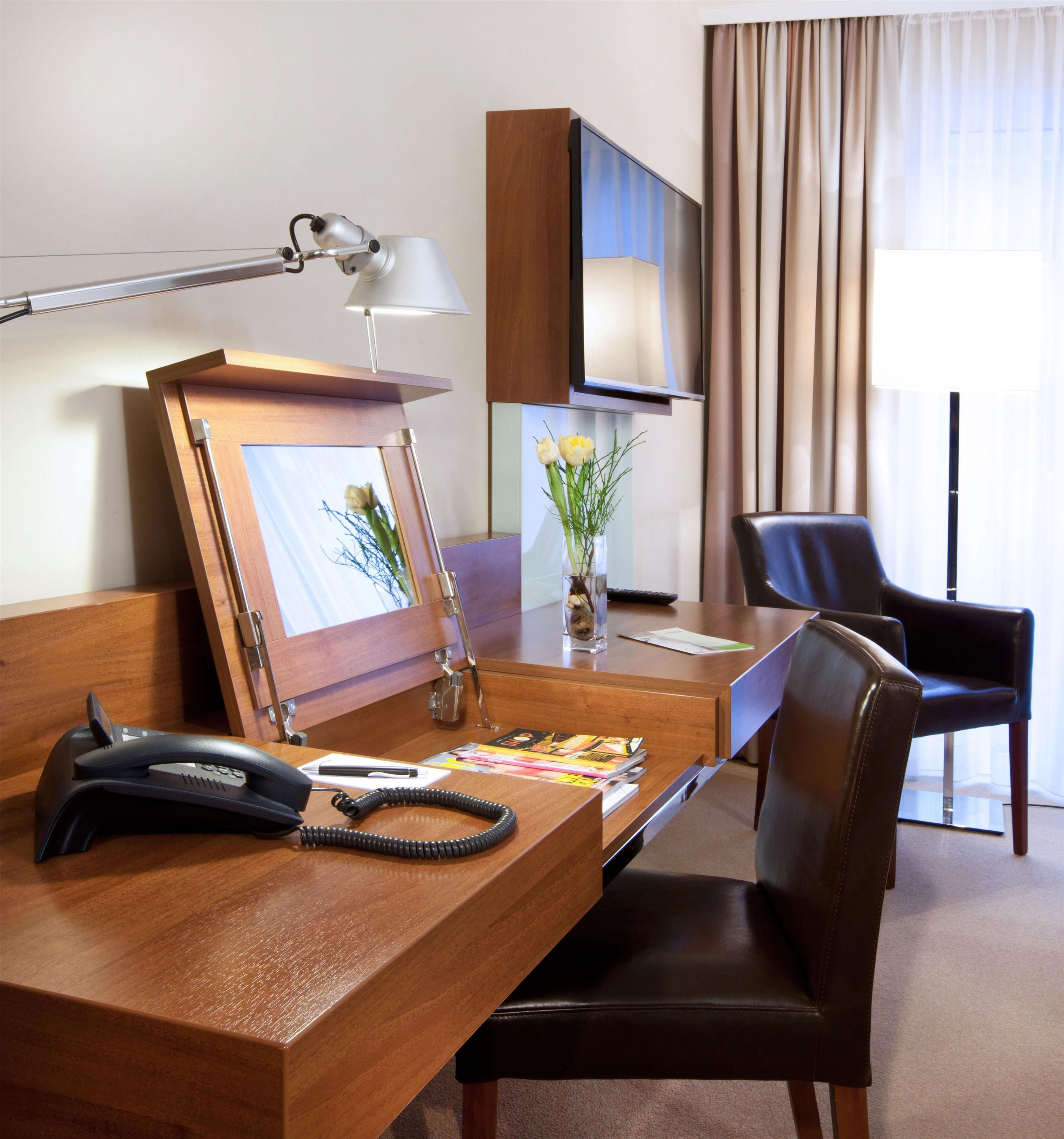 Quarto de hotel com escrivaninha de madeira, telefone, quadro emoldurado, vaso com flores, cadeira e televisão.