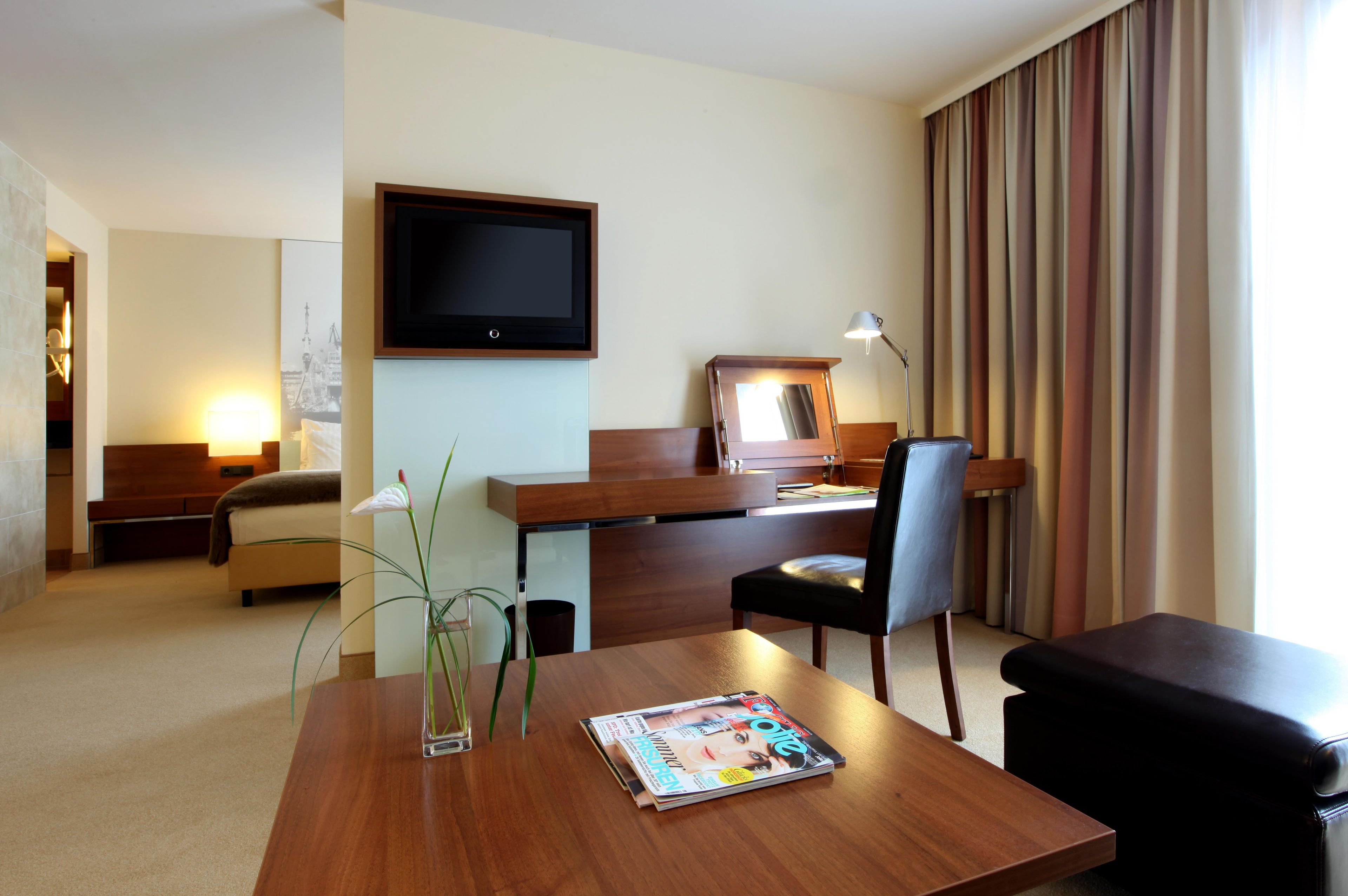Une chambre d’hôtel avec un bureau en bois, un téléviseur fixé au mur, un lit avec une tête de lit, une table de chevet avec une lampe et une fenêtre avec des rideaux.
