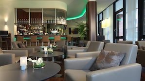 A modern bar area with a curved counter, multiple seating options including sofas and round tables, and a large window providing natural light. - Lindner Hotel Hamburg Am Michel, part of JdV by Hyatt (Hamburg)