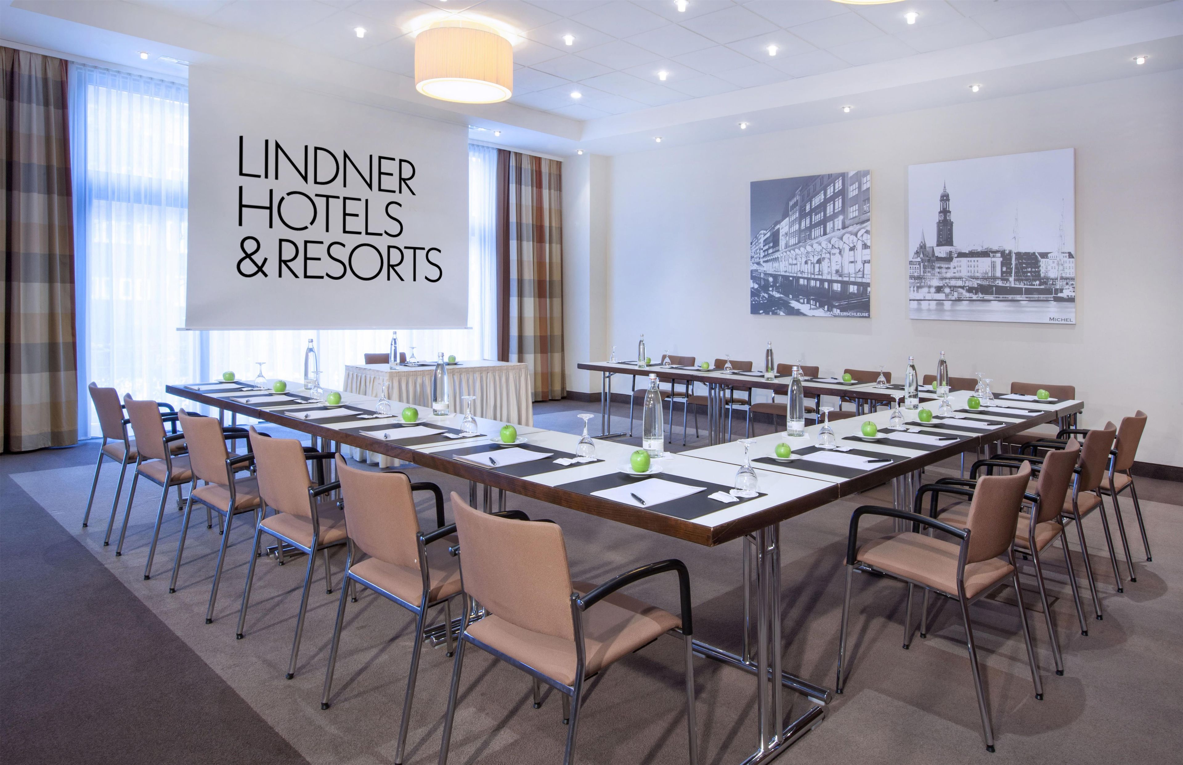 Et mødelokale med et langt bord, stole og et projektorskærm, der viser 'LINDNER HOTELS & RESORTS'.