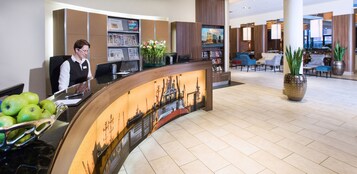Ein Hotel-Empfangsbereich mit einer gebogenen Theke, einer Rezeptionistin, einer Obstschale, einer Blumenvase und einem Stadtbild-Wandgemälde.