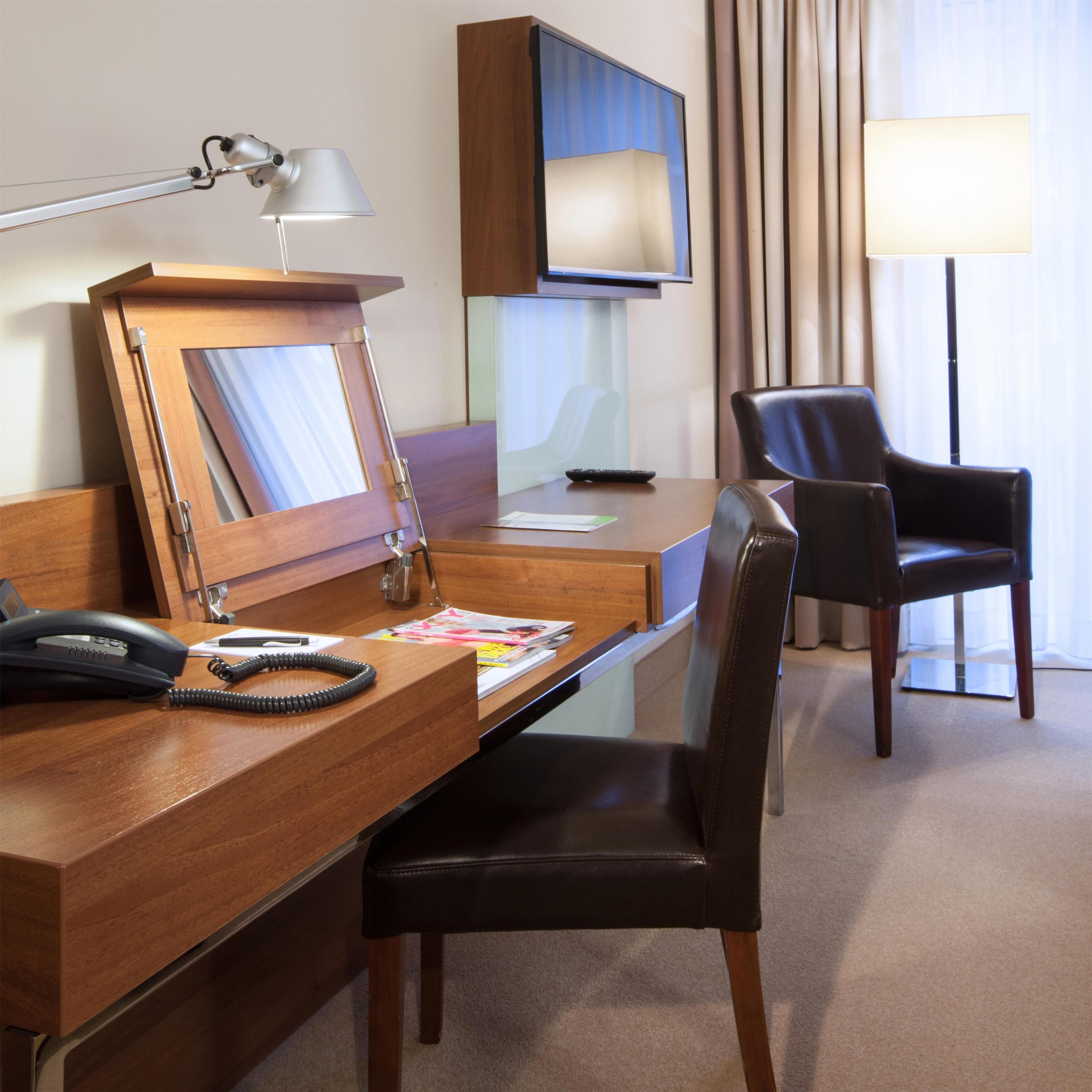 Una habitación de hotel con un escritorio de madera, un teléfono, una revista, un espejo, un televisor y una silla.