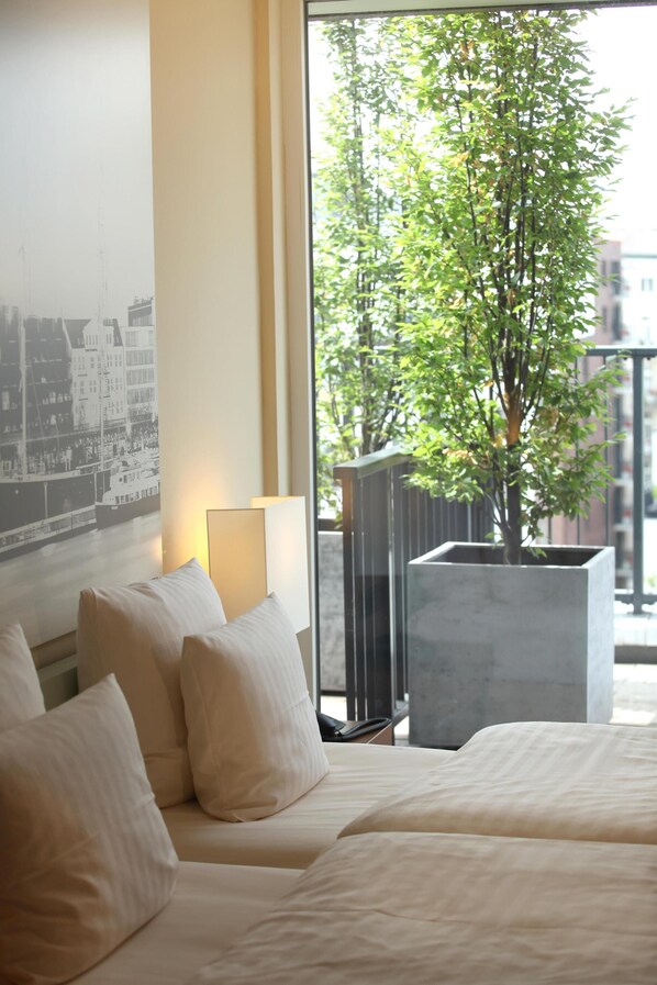 A hotel room with a bed, a large window with a view of a tree, a wall-mounted lamp, and a framed picture.