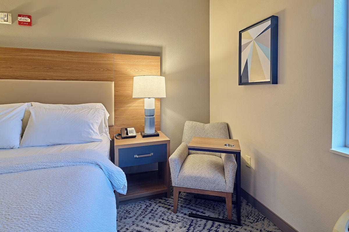 Quarto de hotel com uma cama, mesa de cabeceira com abajur, cadeira e um quadro na parede.
