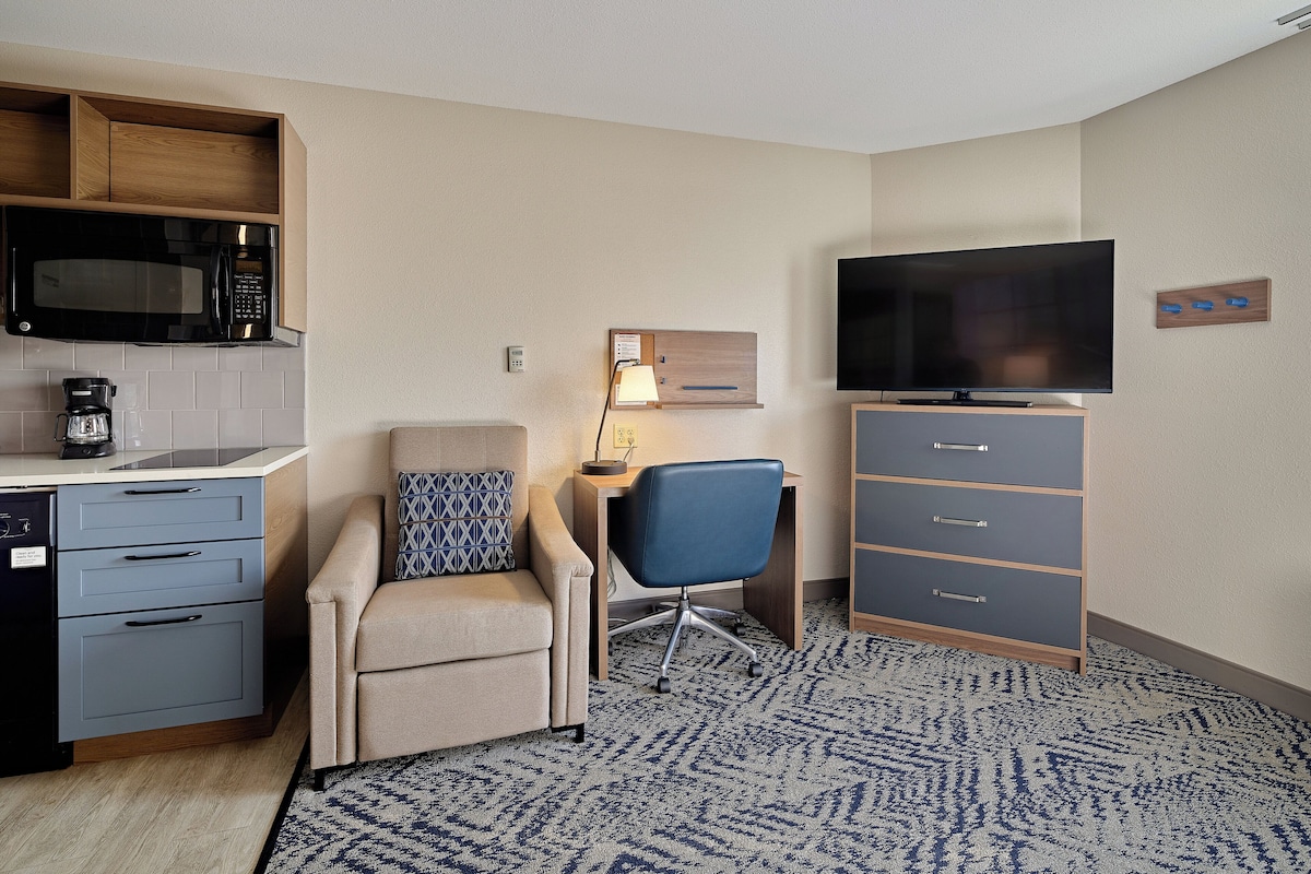 Quarto de hotel compacto com cozinha compacta, sofá, escrivaninha com cadeira, televisão e carpete estampado.
