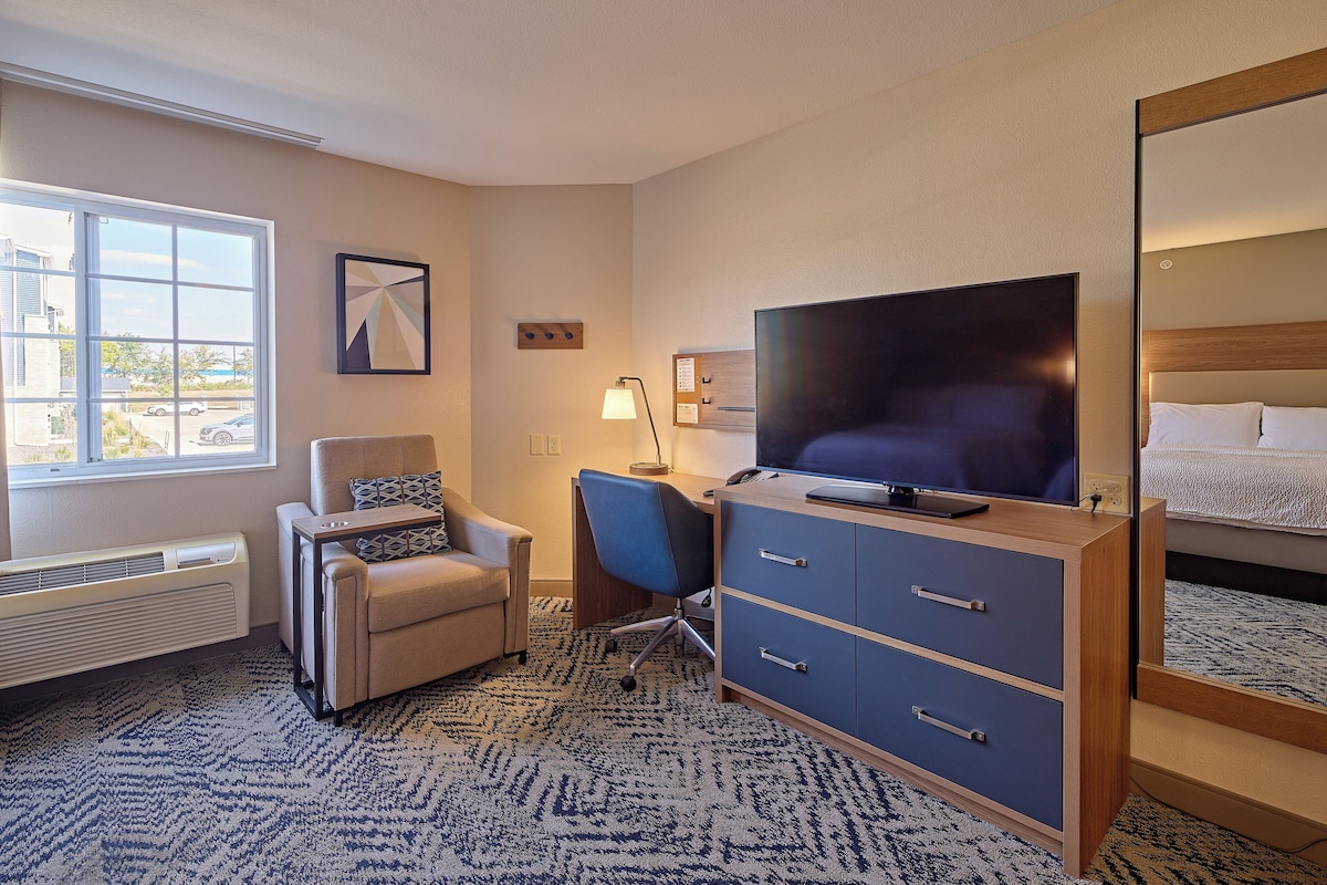 Quarto de hotel com uma janela grande, uma escrivaninha com cadeira, televisão, cama e espelho.