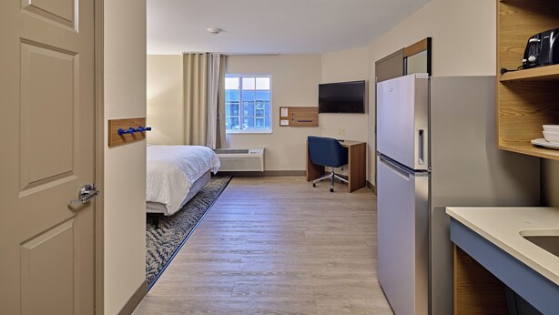 A hotel room with a bed, desk, refrigerator, and a view of the city through the window. - Candlewood Suites Grand Prairie by IHG (Peoria)