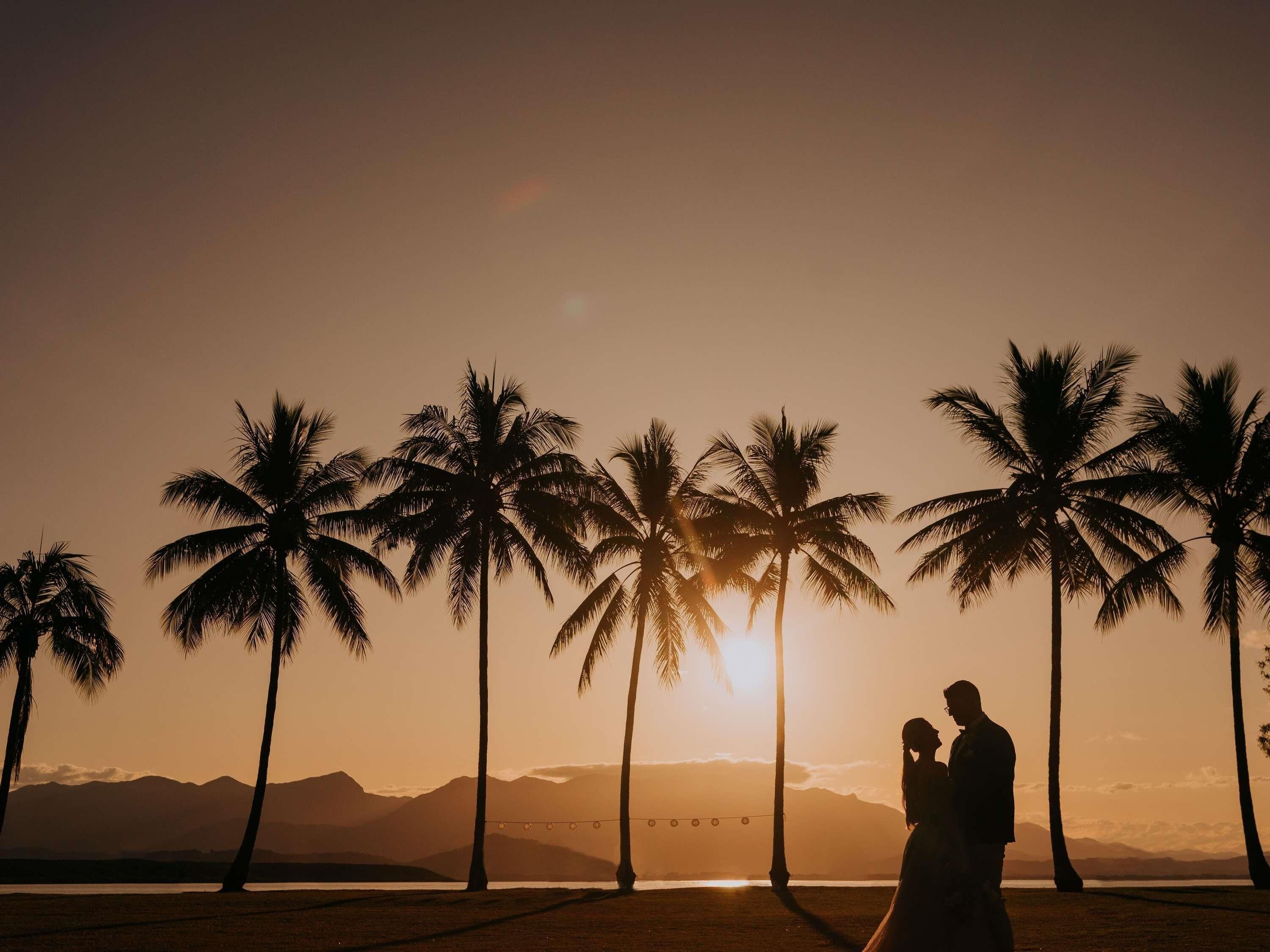 La silhouette d’un couple se détache sur le ciel d’un coucher de soleil, avec des palmiers et des montagnes en arrière-plan.