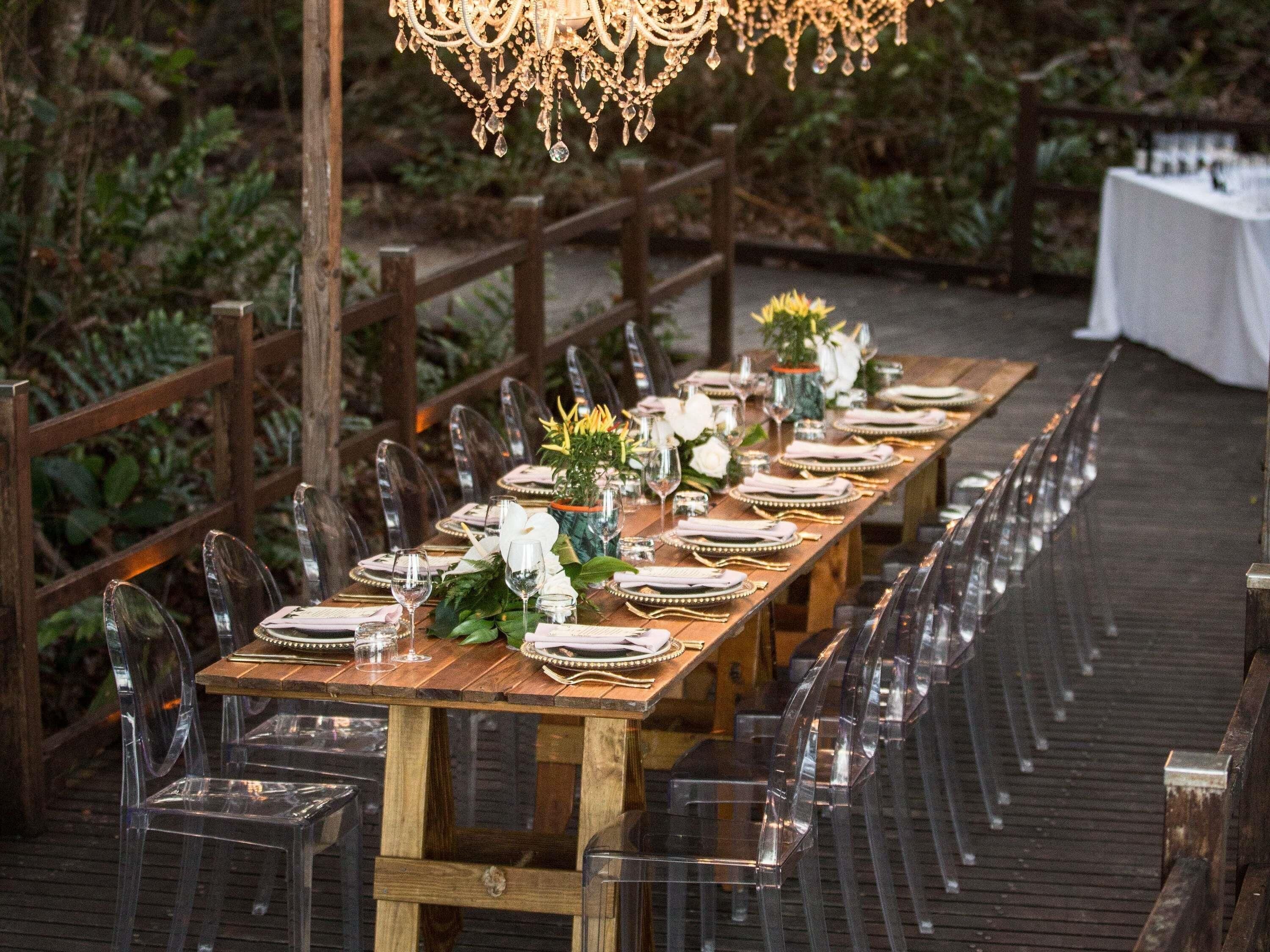 A long wooden table set for a formal outdoor dining event with clear chairs, white tablecloths, and floral centerpieces.
