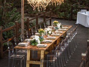 Ein langer Holztisch, gedeckt für ein festliches Dinner im Freien, mit klaren Stühlen, weißen Tischdecken und floralen Tischdekorationen.