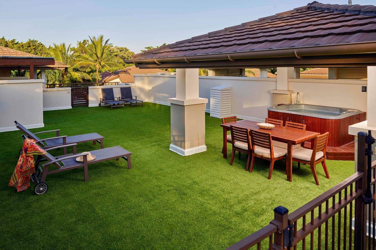 Un jardin avec une pelouse artificielle, un jacuzzi, une table à manger avec des chaises et un espace barbecue couvert.