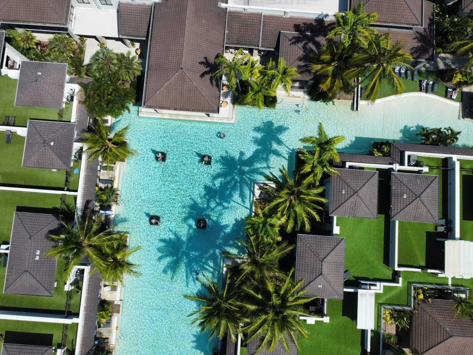 An aerial view of a resort with a central swimming pool surrounded by palm trees and multiple bungalow-style buildings.