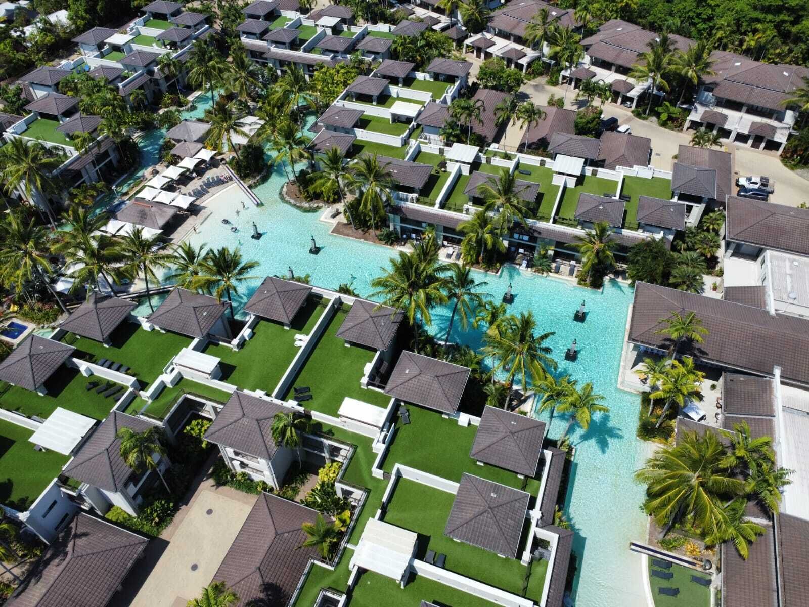 Eine Vogelperspektive zeigt ein Resort mit mehreren bungalowsartigen Unterkünften, einem zentralen Schwimmbecken und umliegenden Palmen.