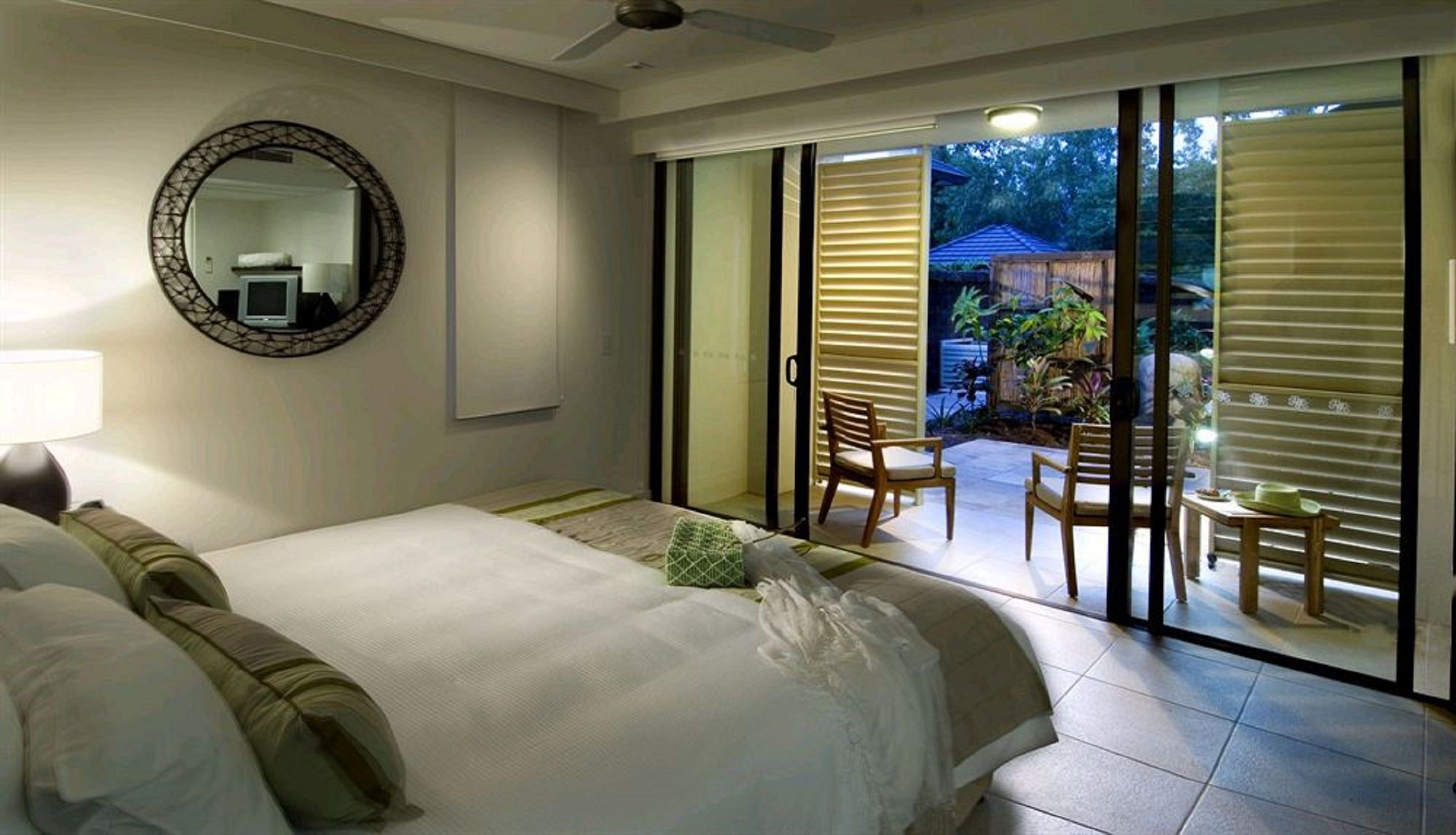 A hotel room with a large bed, a round mirror, and a sliding glass door leading to an outdoor seating area with chairs and a table.