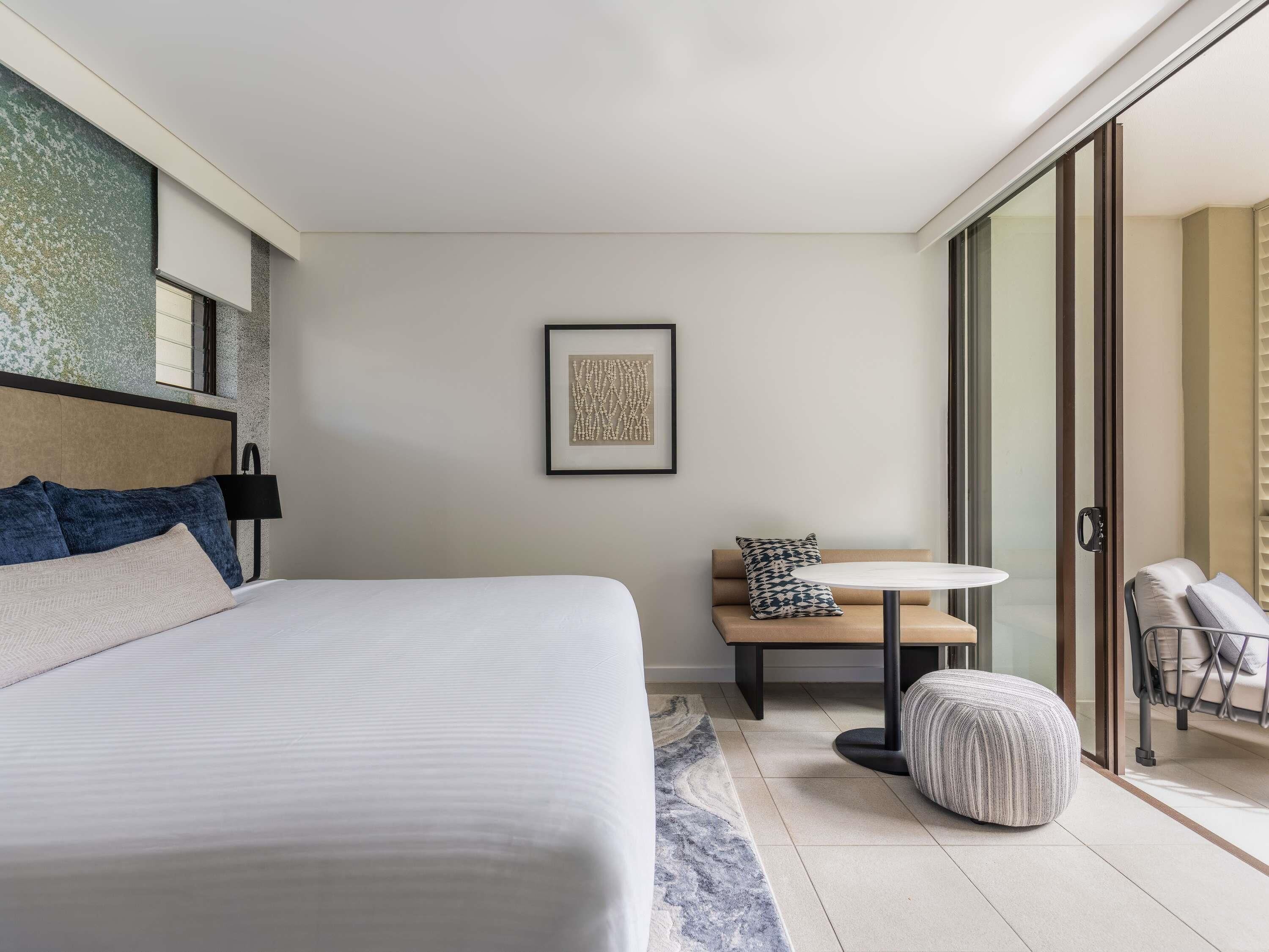 Una camera d'hotel moderna con un letto grande, un tavolino rotondo, una panca e una porta scorrevole in vetro che dà su un balcone.