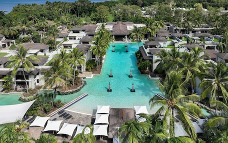 A resort with a central swimming pool, surrounded by villas and palm trees, with a view of the ocean and mountains in the background.