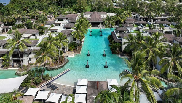 A resort with a central swimming pool, surrounded by villas and palm trees, with a view of the ocean and mountains in the background.