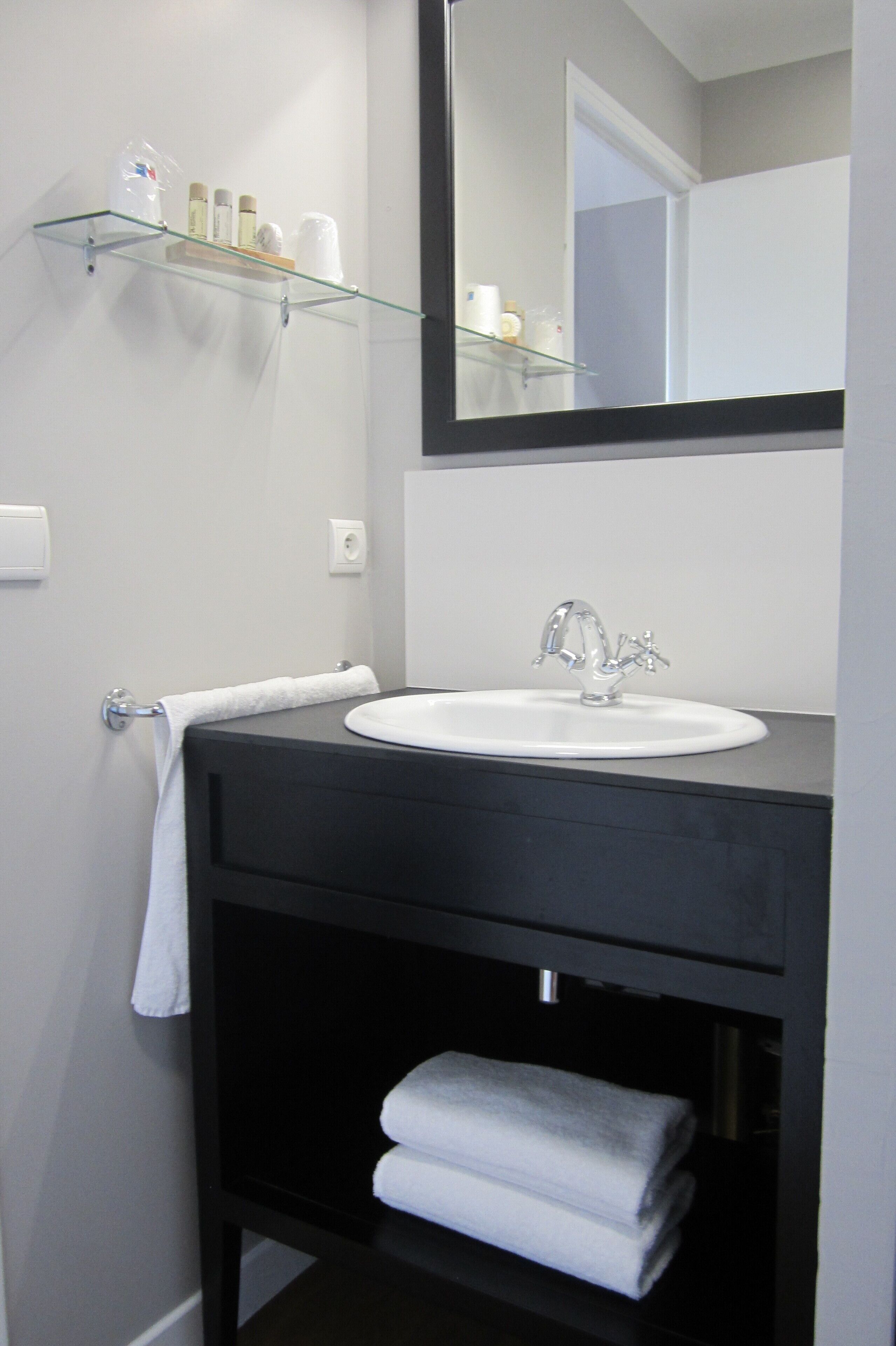 Un baño moderno con un mueble de tocador negro, lavamanos blanco y un estante de vidrio sobre el lavamanos con artículos de aseo.