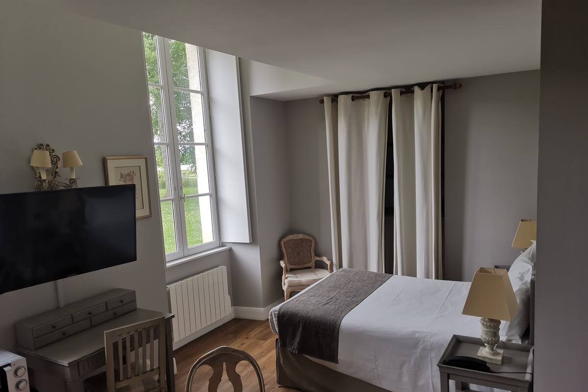 A hotel room with a bed, a desk with a computer, a chair, a TV, and a window with curtains.