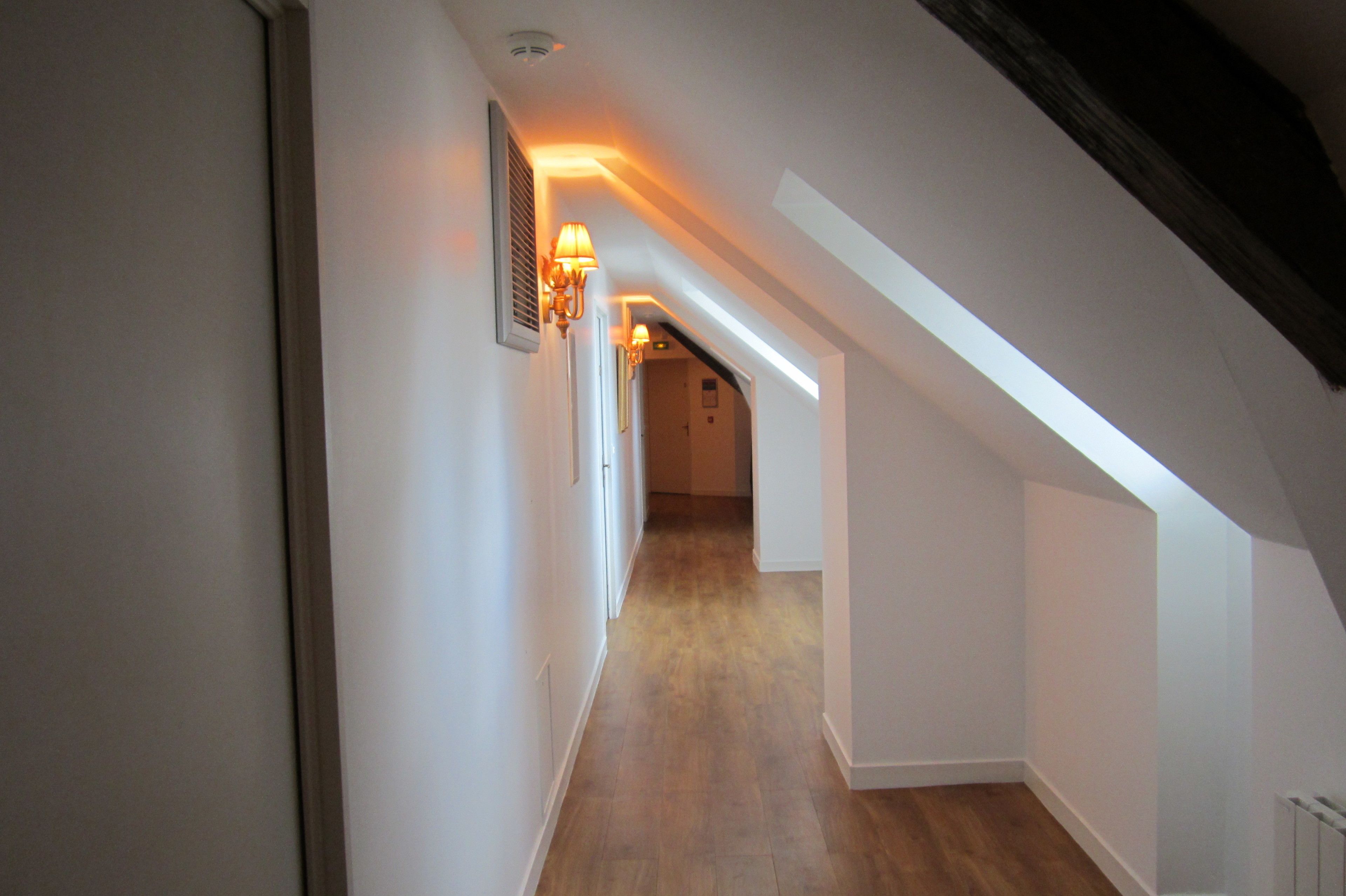 Un pasillo estrecho con suelo de madera y paredes blancas, que conduce a una habitación con techo inclinado y un espacio de ático visible.