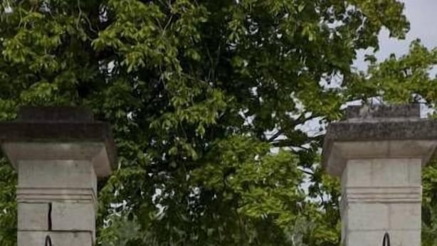 Un edificio con fachada simétrica, que presenta una entrada central flanqueada por dos columnas con faroles. Ventanas distribuidas uniformemente a lo largo de la fachada, y una pequeña placa es visible en la columna izquierda.