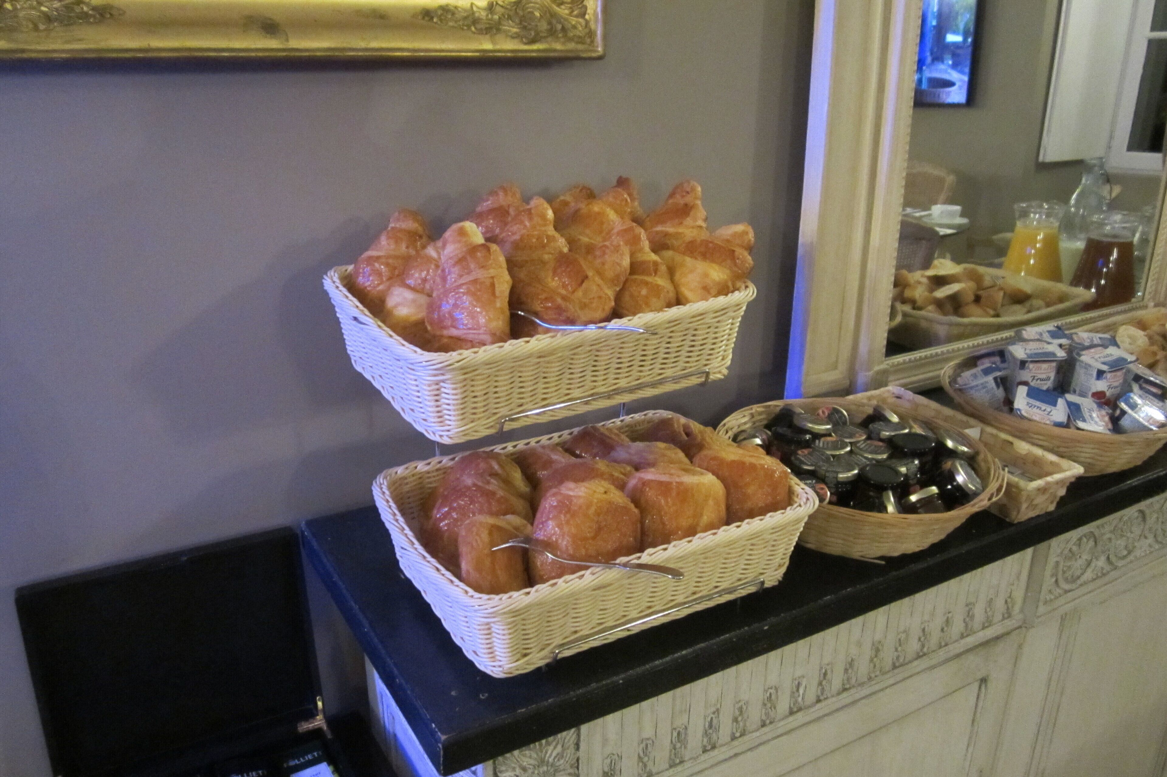 A buffet counter with baskets of croissants and other pastries, a selection of beverages, and a mirror reflecting the setup.