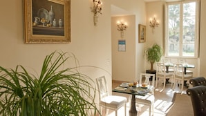 Comedor con una mesa dispuesta para cuatro personas, rodeada de sillas blancas. En primer plano, hay una planta verde grande, y un cuadro sobre la mesa.