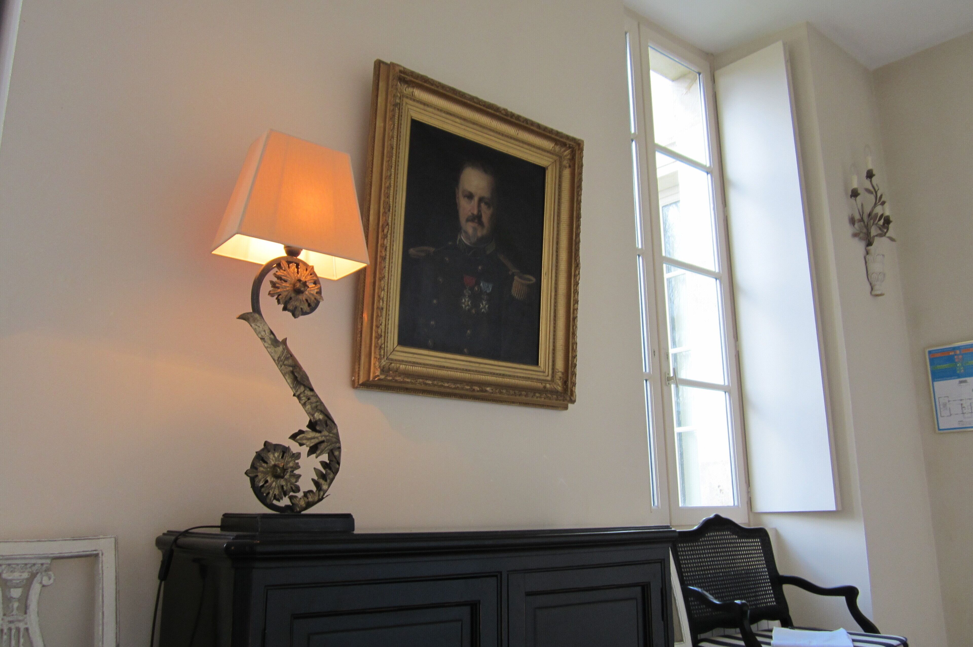 Ein Zimmer mit einem gerahmten Porträt an der Wand, einer Lampe, einem Sessel und einem Fenster.