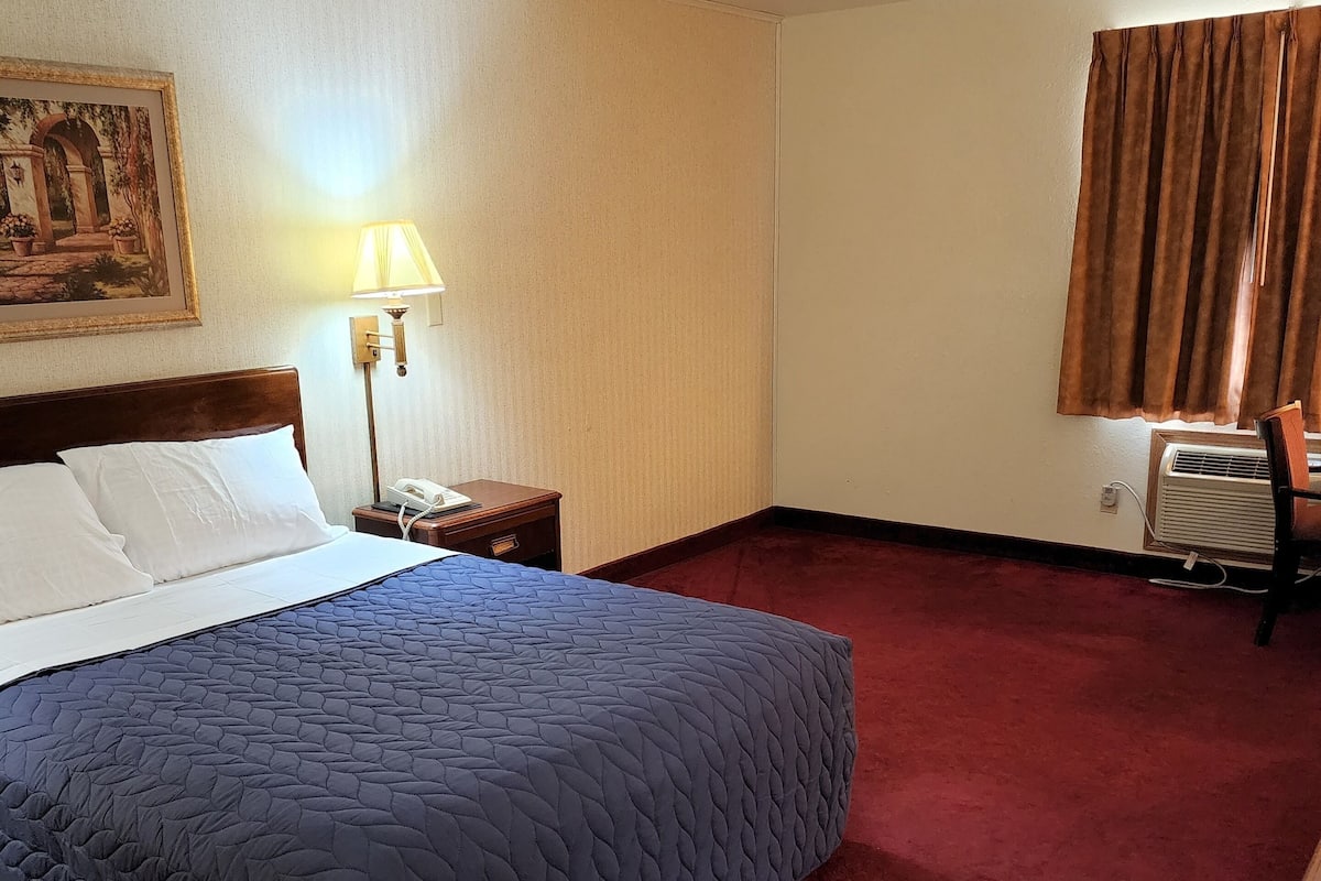 A hotel room with a large bed, a nightstand, a lamp, a painting on the wall, and a window with curtains.
