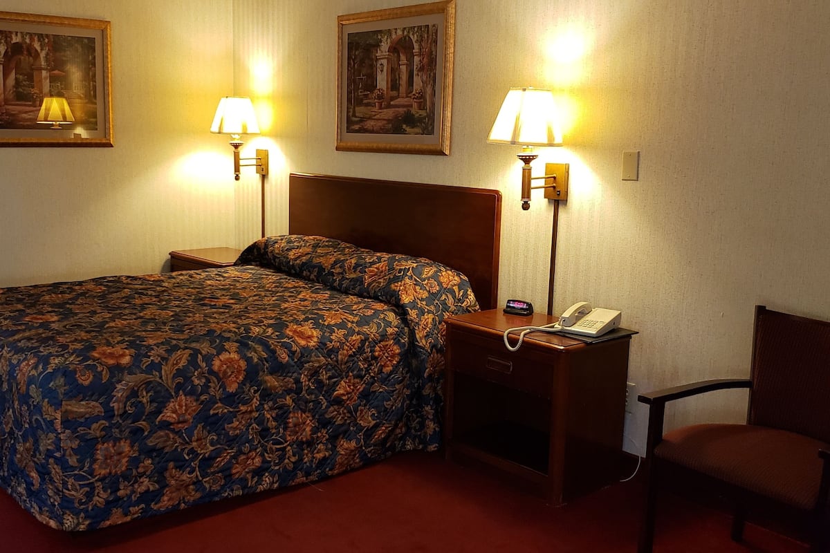 A hotel room with a large bed, two bedside lamps, a nightstand with a phone, and a chair.