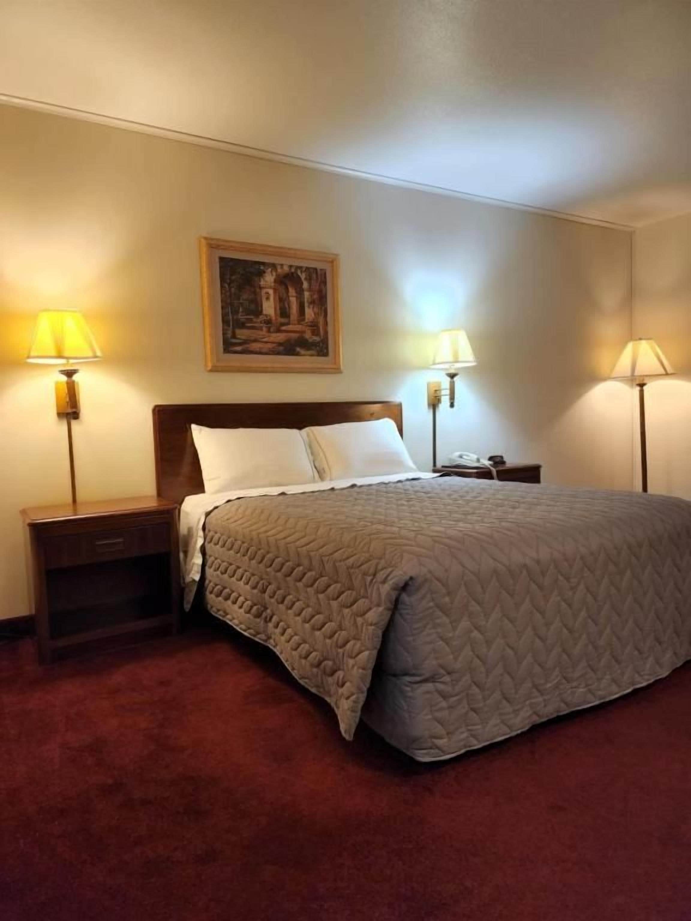A hotel room with a bed, two bedside lamps, a nightstand, and a painting on the wall.
