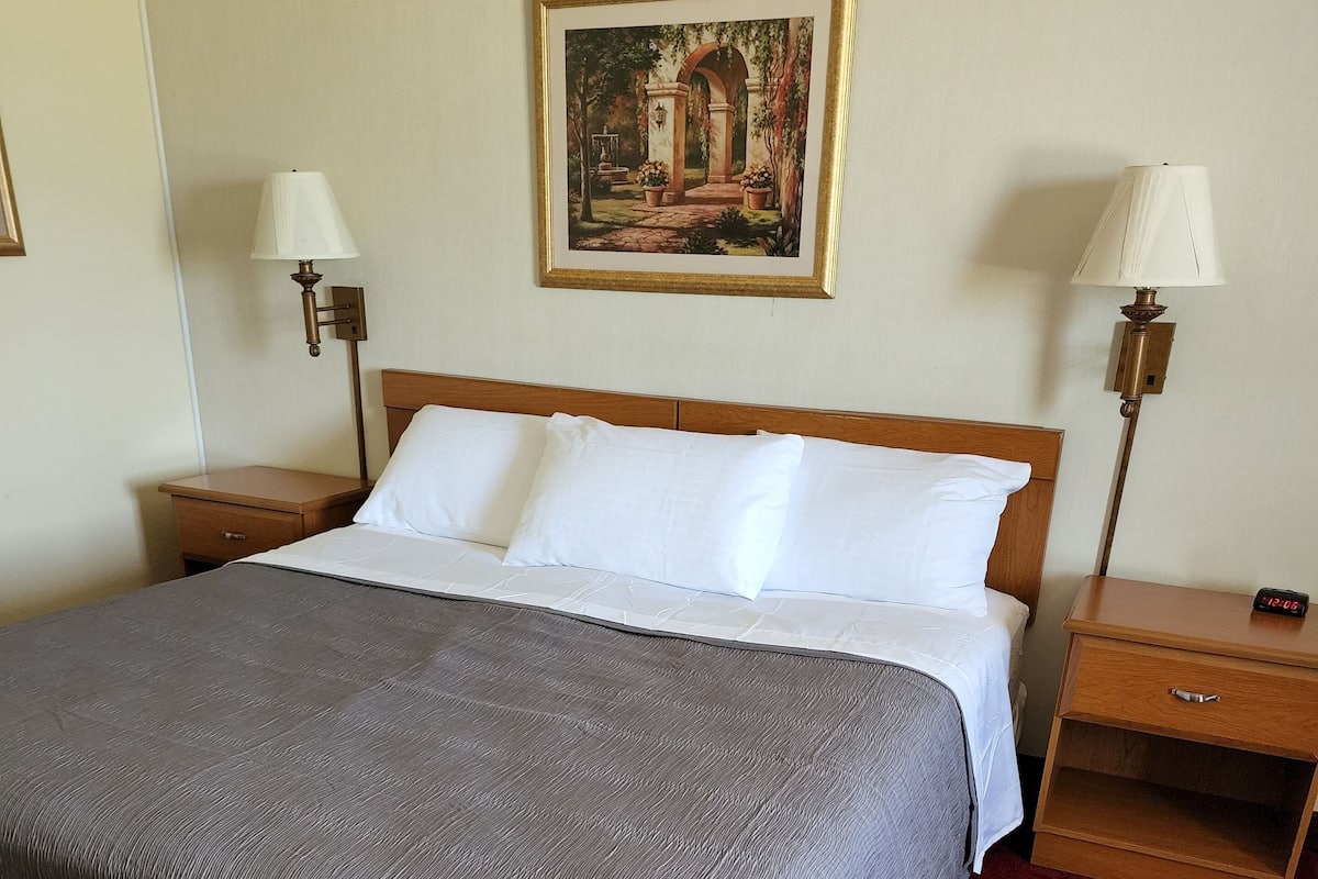 A neatly made bed with a grey blanket, two bedside tables with lamps, a wooden nightstand, a small drawer, and a framed picture on the wall.
