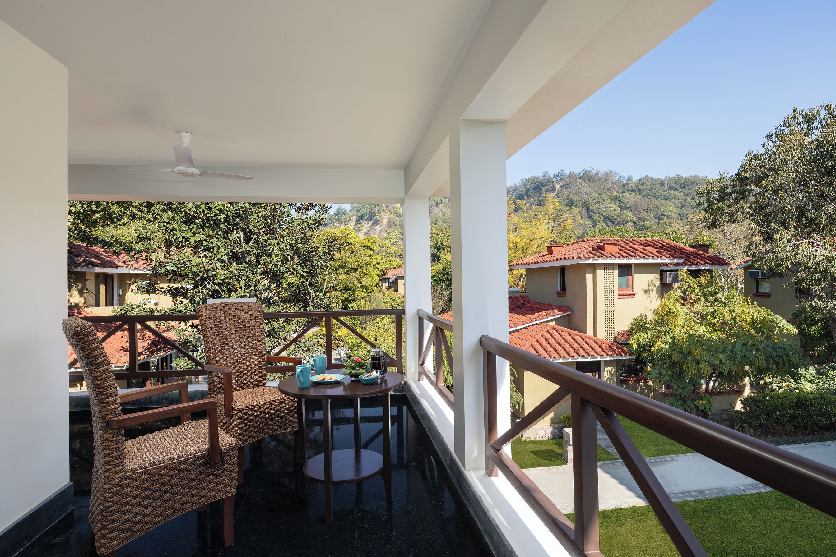 Ein Balkon mit Korbstühlen und einem Tisch, mit Blick auf eine üppige grüne Landschaft und angrenzende Häuser.