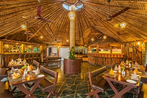 Ein Restaurant mit einem hohen, reetgedeckten Dach und einer zentralen Säule. Tische sind gedeckt mit Servietten, Besteck und Gläsern. Im Hintergrund ist eine Bar zu sehen.