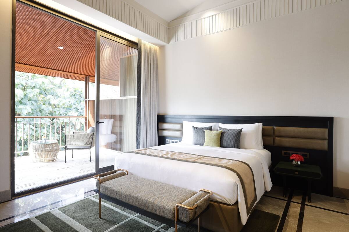 A modern hotel room with a large bed, a small table with a vase of flowers, a bench, and a balcony with wicker furniture.