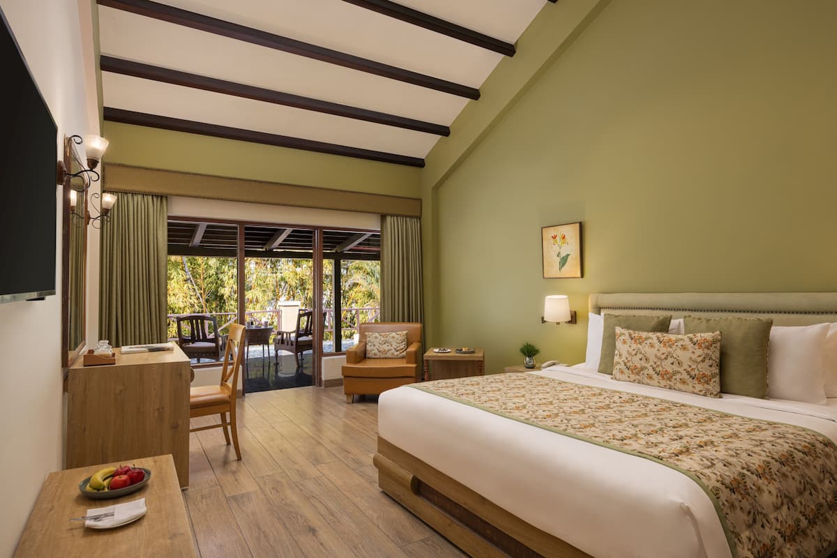 A hotel room with a large bed, a desk with a chair, a dining table with fruit, and a balcony with a chair.