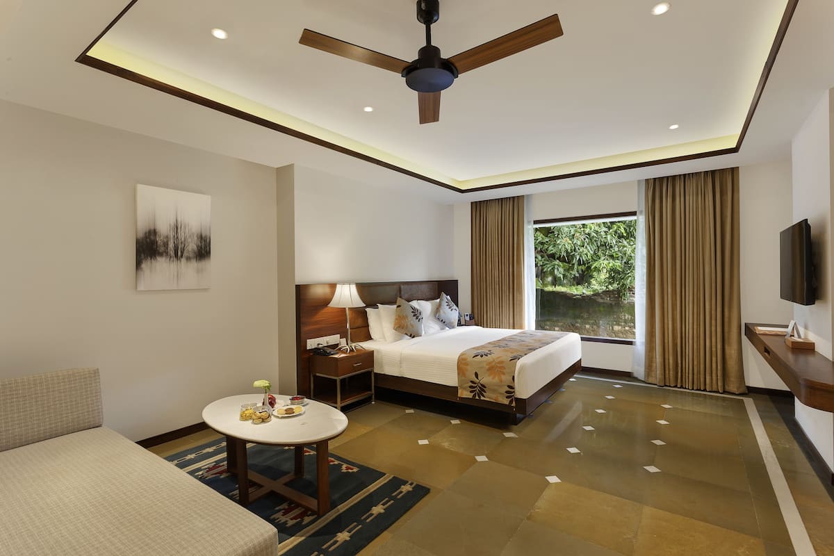 A hotel room with a bed, bedside table, lamp, and a view of greenery outside.
