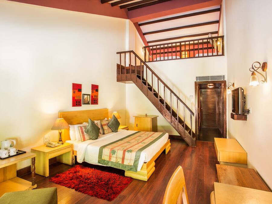 A hotel room with a wooden staircase, a bed with a red rug, a wooden nightstand, and a desk with a lamp.