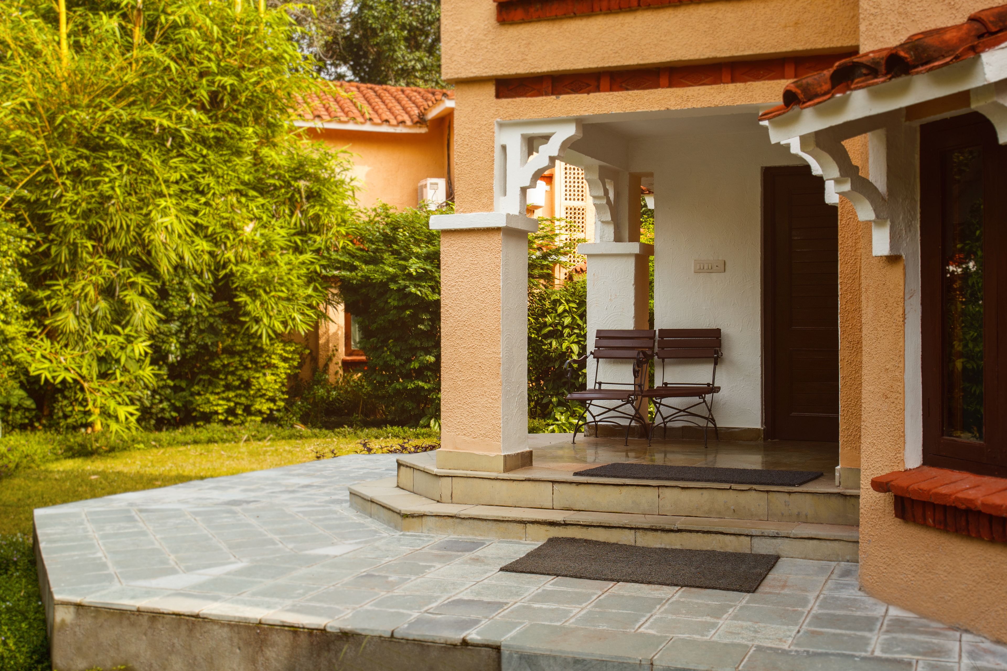 A covered outdoor seating area with a bench and a table, surrounded by greenery and a stone-paved entrance.