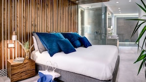 A modern bedroom with a wooden slat wall, a large bed with blue pillows, a bedside table with a magazine, and a bathroom visible through a glass door.